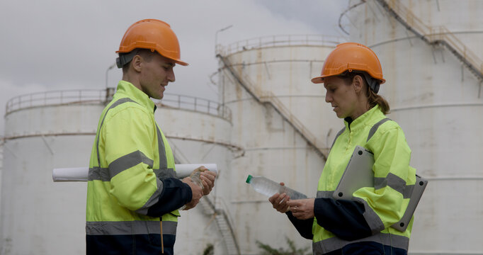 Male and female energy engineers collaborate with a laptop, blueprint, and digital tablet at the oil storage tanks in chemical and refinery  plant