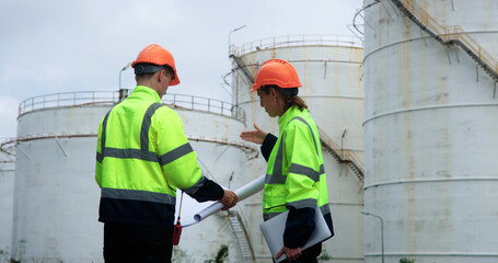 Male and female energy engineers collaborate with a laptop, blueprint, and digital tablet at the oil storage tanks in chemical and refinery  plant