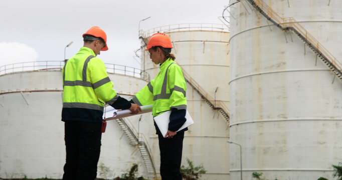 Male and female energy engineers collaborate with a laptop, blueprint, and digital tablet at the oil storage tanks in chemical and refinery  plant