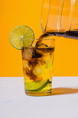 Coffee being poured on glass of lemonade with slice of lime on vibrant yellow background