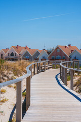 Fototapeta premium A scenic wooden boardwalk through sandy dunes leads to vibrant red and white striped houses in Costa Nova, Aveiro, Portugal, under a clear blue sky.