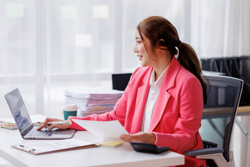 Young attractive business asian woman working of reading paper accounting file financial report, tax invoice on her documents project with laptop computer in the workplace office.