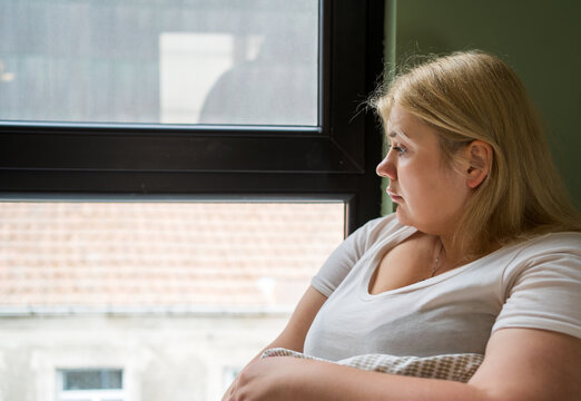 Plus size woman sitting by window, looking out with thoughtful expression. Reflecting on life, possibly experiencing loneliness, depression, or heartbreak.