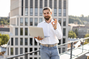 Confident business man standing on rooftop working on laptop making okay hand sign expressing success positivity with urban background in daylight