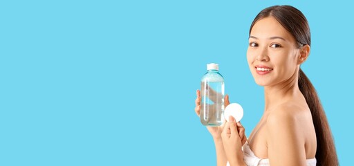 Young Asian woman holding micellar water and cotton pad on blue background with space for text