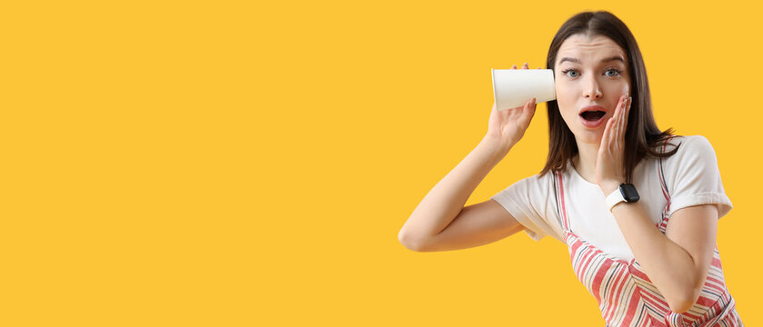 Shocked young gossip woman with cup trying to hear something on yellow background