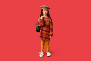 Cute little girl in stylish clothes with backpack on red background