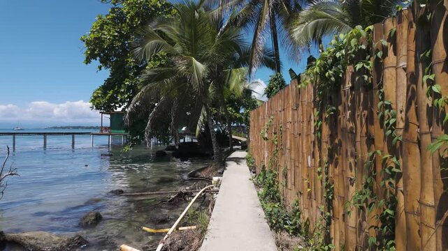 Point of view walking along a coastal path beside a fence on Bastimentos Island, Panama. Tropical scenery with lush greenery and calm waters.