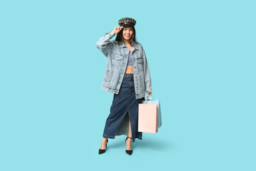 Fashionable young Asian woman in denim clothes with shopping bags on blue background