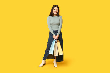 Fashionable young Asian woman in stylish denim skirt with shopping bags on yellow background