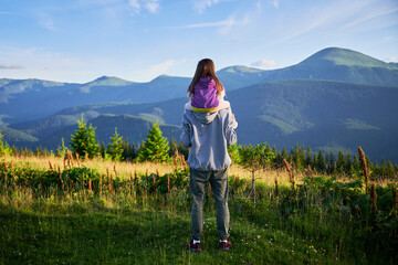 Father carries his young daughter on his shoulders, both enjoying scenic mountain view. They surrounded by lush greenery and rolling hills under clear blue sky.
