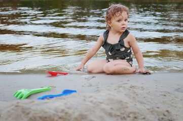 Curly haired baby girl dressed in polka-dot swimsuit enjoying playtime with toys on the coast playing with a shovel on the beach enjoying summer holiday vacation