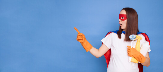 Astonished shocked woman posing as superhero with mask and cape holding detergent spray and brush performing household tasks isolated over blue background pointing at advertisement area © sementsova321