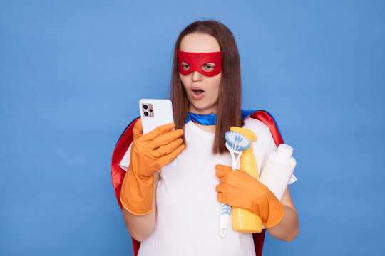 Shocked astonished woman in housemaid uniform with cell phone and detergent spray browsing internet for cleaning tips on mobile application isolated over blue background