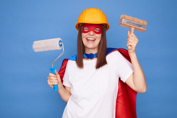 Professional woman in superhero costume with paintbrush and roller preparing for home renovation project dancing rejoicing good start of working day isolated over blue background