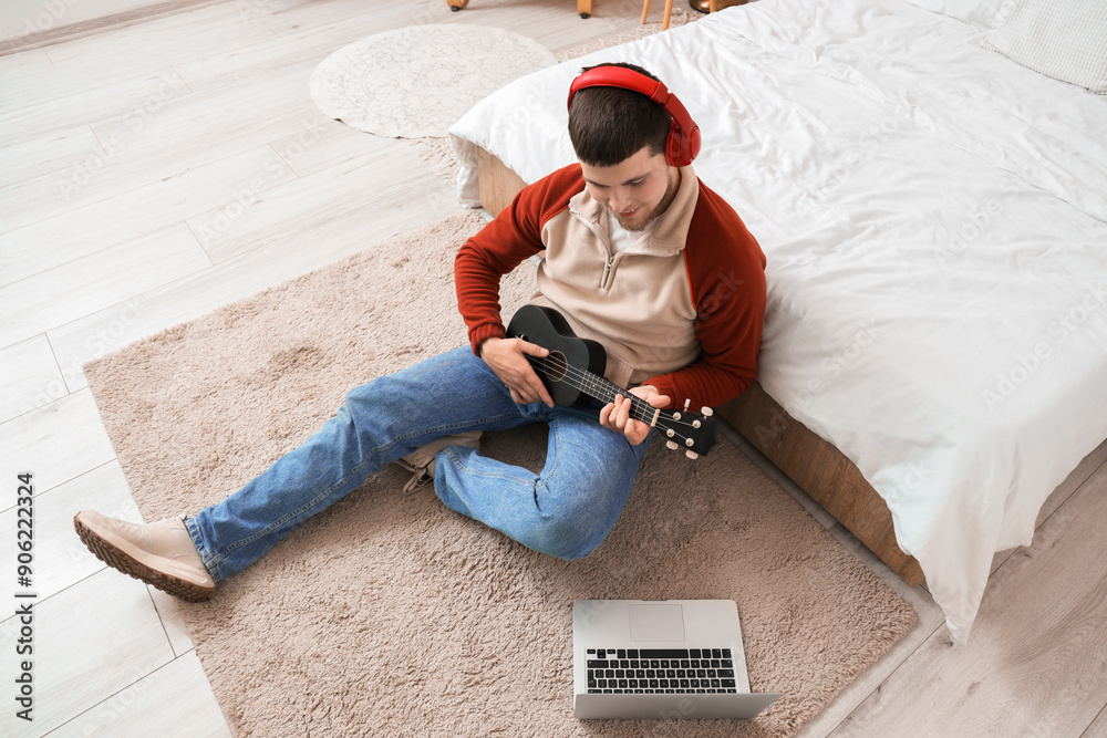 Wall mural young bearded man with headphones playing ukulele in bedroom