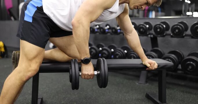 Young man performs dumbbell row exercise in modern gym slow motion. Athlete improves upper body enhancing posture during workout in club