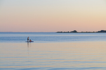 Fototapeta premium Paddleboarders navigate calm waters as the sun sets, painting the sky with gentle pastel hues, creating a serene evening moment.