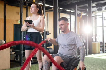 Side view of man with disability using wheelchair during strength training in gym with female coach...