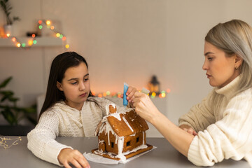 mother and daughter decorating gingerbread house. Beautiful living room with lights. Happy family celebrating holiday together.