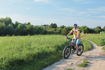 Naklejka premium Cyclist in helmet with equipment on electric bicycle rides along path in field in countryside. Sports and health. Summer hobby. 