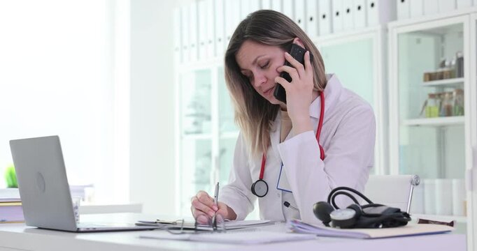 Lady medical specialist speaks on phone making notes on papers. Female doctor consults patient at table from hospital office with deep focus