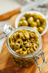 Sliced pickled green olives in brine in jar on wooden cutting board.