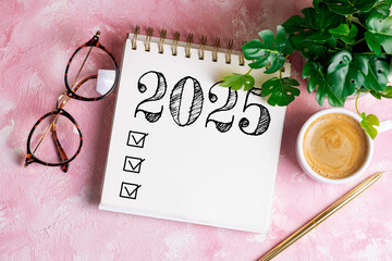 New year resolutions 2025 on desk. 2025 goals list with notebook, coffee cup, plant on pink table....