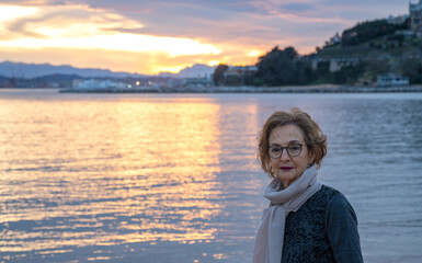 Elderly woman wearing glasses and a scarf, standing by the water at sunset