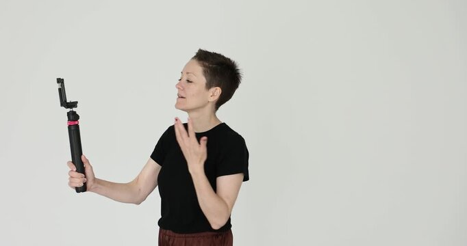 Woman with active facial expression records video on phone using monopod on white background. Female blogger creates content with equipment
