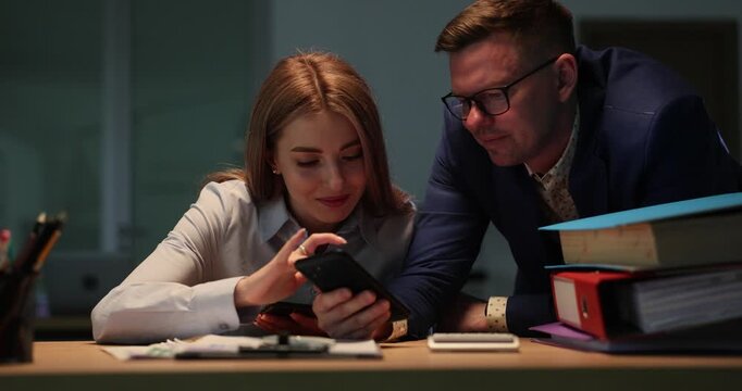 Man colleague holds smartphone and woman reads email from customer at workplace in office. Responsible workers solve work issues in evening