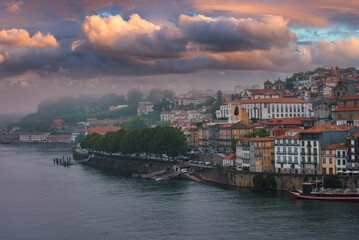 Obraz premium A stunning view of Porto, Portugal, featuring the Douro River, vibrant buildings with red-tiled roofs, a prominent white church, and lush greenery under a dramatic sky.