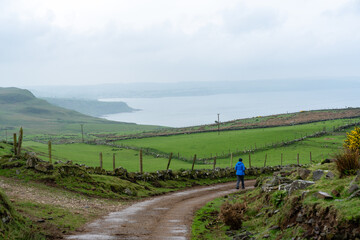 Frau in blauer Jacke und Mütze wanders durch die Landschaft zwischen Feldern und Wiesen von...