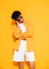 Young attractive man with short curly hair hairstyle. Smiling handsome male in casual stylish yellow shirt and shorts clothes posing in studio, isolated. Cheerful and happy model, sunglasses