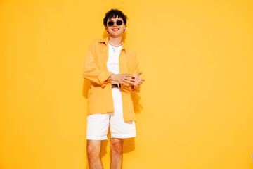 Young attractive man with short curly hair hairstyle. Smiling handsome male in casual stylish yellow shirt and shorts clothes posing in studio, isolated. Cheerful and happy model, sunglasses