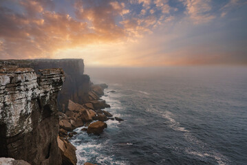 Breathtaking view of the western coast of Portugal featuring dramatic cliffs, vibrant sunset hues, and misty ocean spray. Waves crash against the rugged rocks.