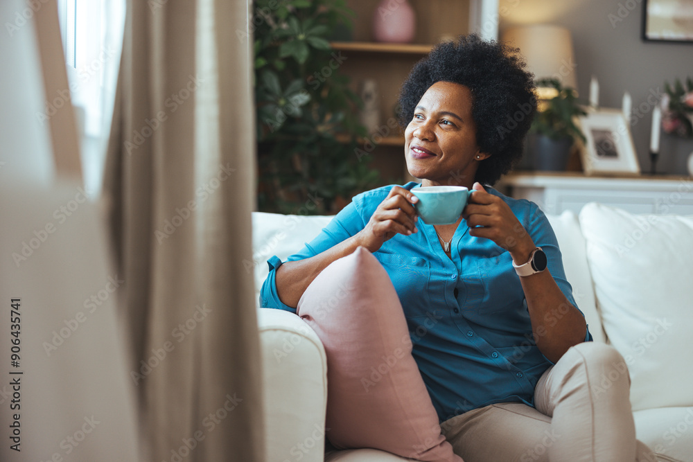 Wall mural happy young african woman sitting on comfortable sofa in living room at home. smiling pretty girl dr