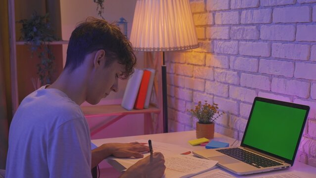 Focused adult man college student sitting at desk in apartment room late at night doing homework with laptop chroma key green screen, writing essay. Education concept.