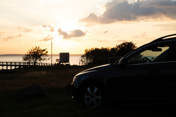 Fototapeta premium Car camping in Skåne south of Helsingborg. A beautiful midsummer nights sunset at the beach. Light mirroring in freshly polished car. dramatic clouds.