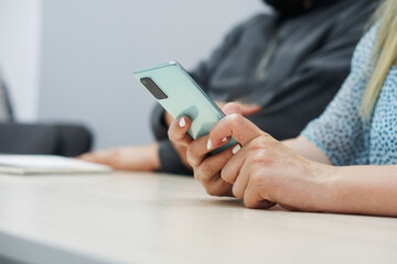 Hands of a woman sitting at a table in an office with a smartphone. Online correspondence, planning and routine. Workspace. Photo. No face. Selective focus