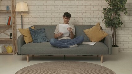 Focused adult man college student sitting on sofa in living room at daylight and doing homework, holding notebook reading essay. Education concept.