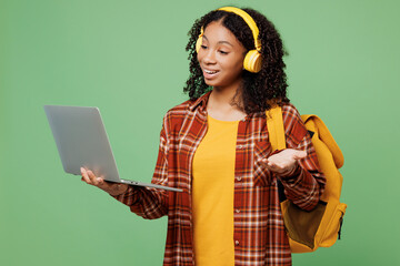 Young fun teen girl IT student wear brown yellow casual clothes headphones backpack bag use work on laptop pc computer talk speak isolated on plain green color background. High school college concept.