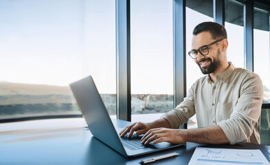 A smiling young man in a suit sits in the office at a desk and works on a laptop, typing on a keyboard, texting, working with data