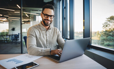 A smiling young man in a suit sits in the office at a desk and works on a laptop, typing on a keyboard, texting, working with data