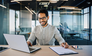 A smiling young man in a suit sits in the office at a desk and works on a laptop, typing on a keyboard, texting, working with data