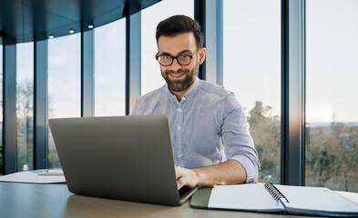 A smiling young man in a suit sits in the office at a desk and works on a laptop, typing on a keyboard, texting, working with data
