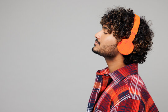 Side profile view young happy smiling Indian man he wears red shirt casual clothes listen to music in headphones close eyes isolated on plain grey color background studio portrait. Lifestyle concept.