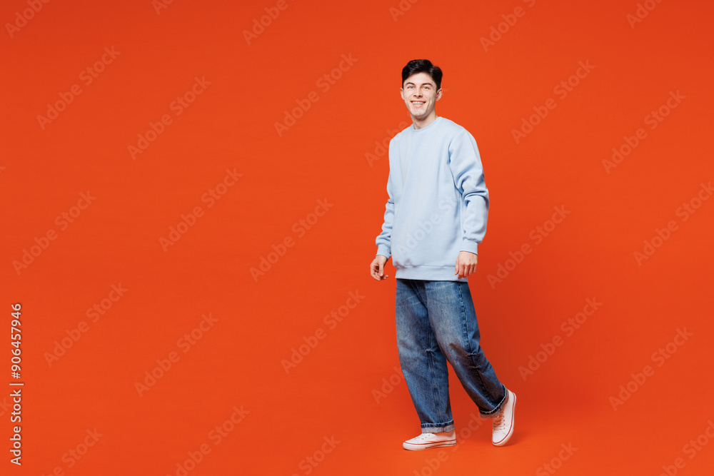 Wall mural Full body side profile view young smiling happy Caucasian man wears blue sweatshirt casual clothes walk go look camera isolated on plain red orange color background studio portrait. Lifestyle concept.