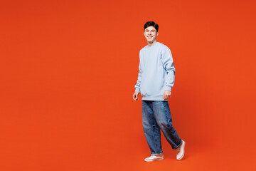 Full body side profile view young smiling happy Caucasian man wears blue sweatshirt casual clothes walk go look camera isolated on plain red orange color background studio portrait. Lifestyle concept.