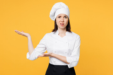 Young surprised indignant chef cook baker woman wear white shirt black apron uniform toque chefs hat point hands arms aside on area isolated on plain yellow background studio. Cooking food concept.